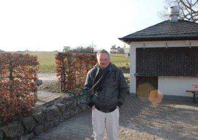 Ein Mann mit Brille und Schal steht vor einem Häuschen und neben braunen Hecken.