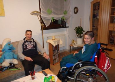 Zwei Männer sitzen in einem Wohnzimmer, die Person auf der rechten Seite sitzt im Rollstuhl. Auf dem Sofa steht eine große Schlumpf-Figur
