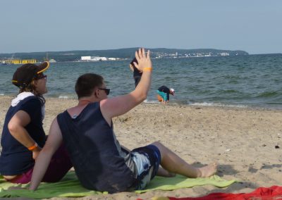 Zwei Personen liegen auf einem Handtuch am Strand. Die rechte Person hebt die Hand.